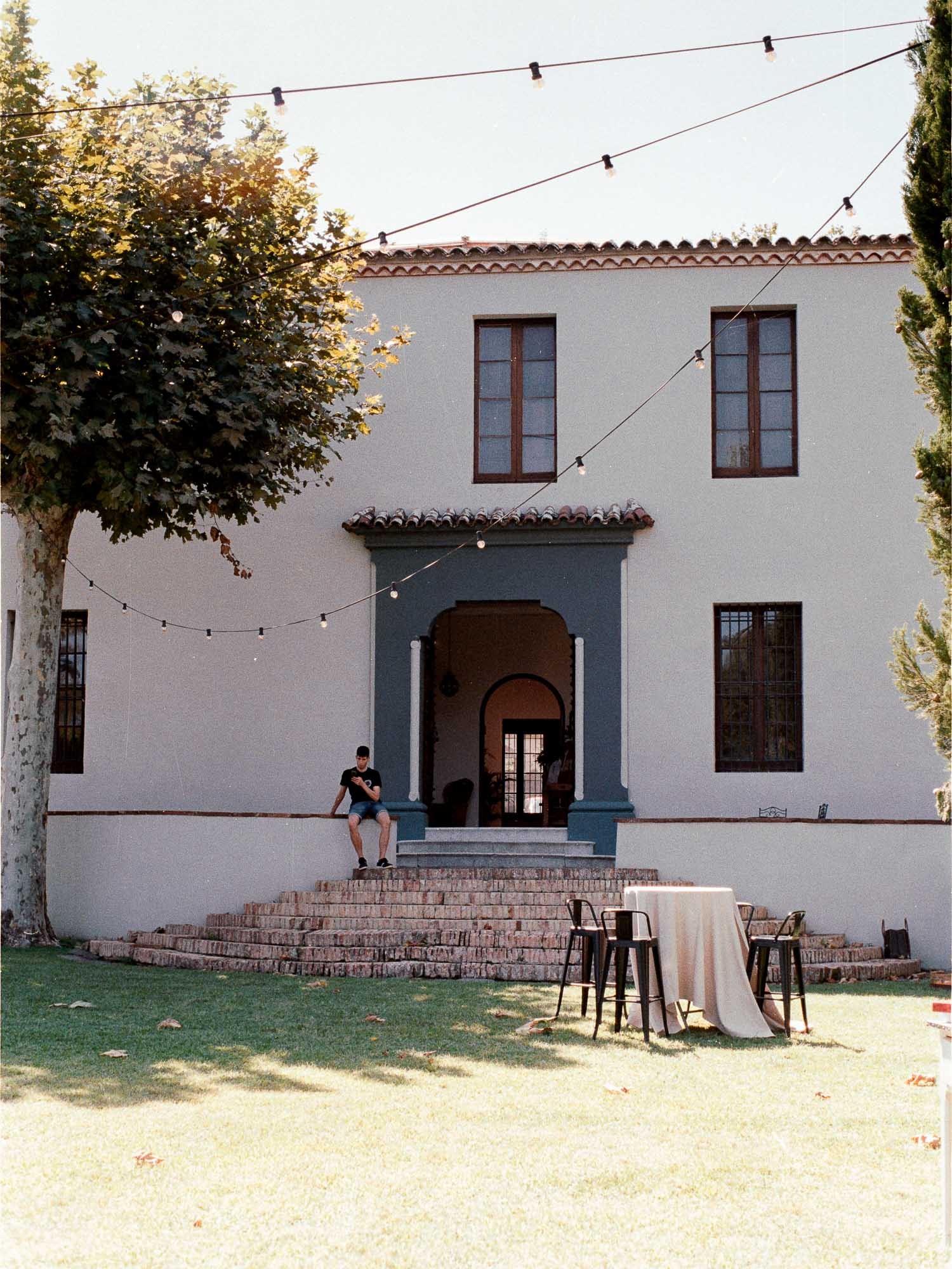 White masia facade with an arched entrance and steps, string lights above and a lawn with a table in sunlight, Anna and Bernat’s wedding at Masia Montcorp
