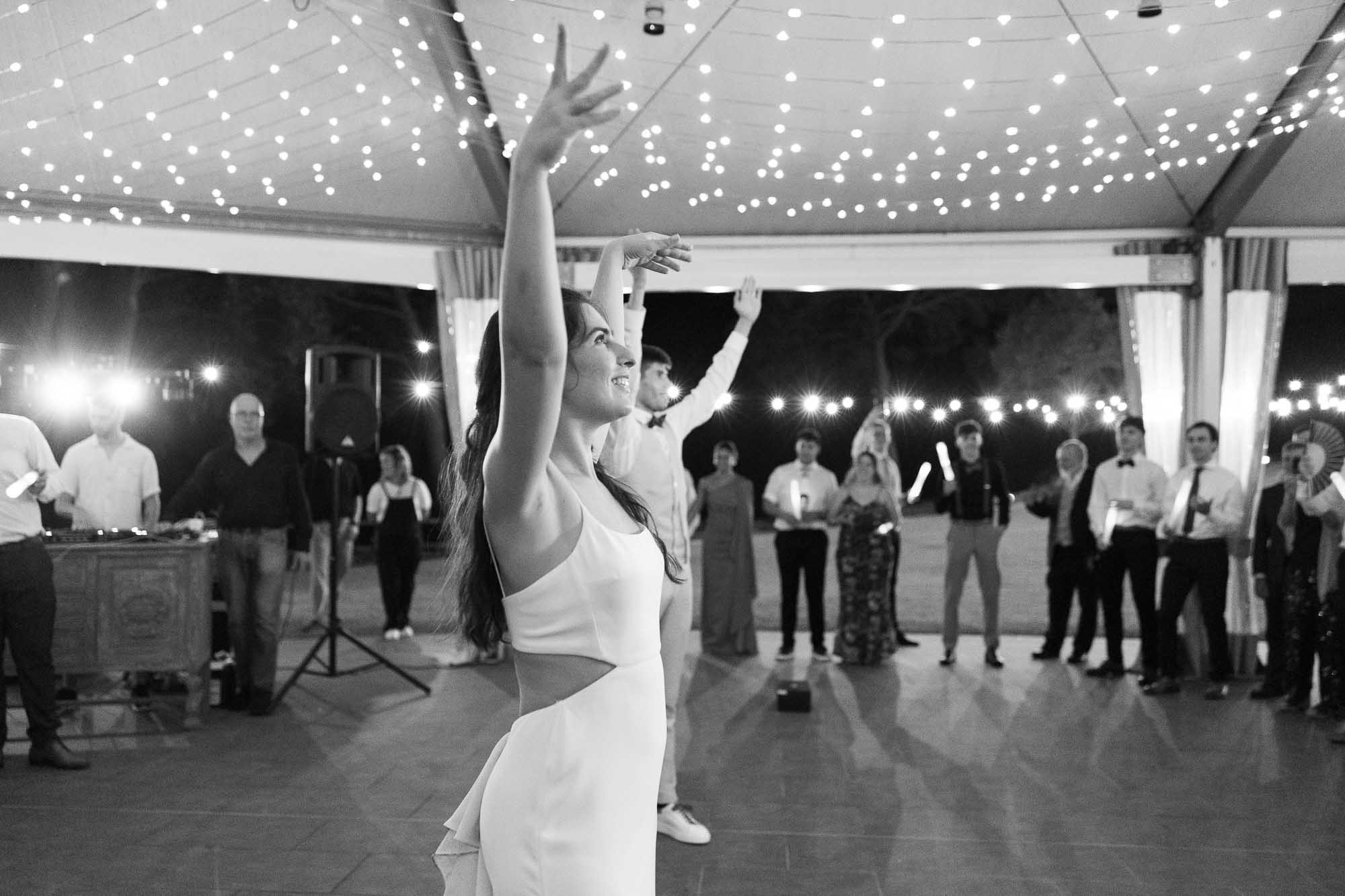 Black and white wide shot of the bride dancing with an arm raised inside a circle of guests under string lights, Anna and Bernat’s wedding at Masia Montcorp