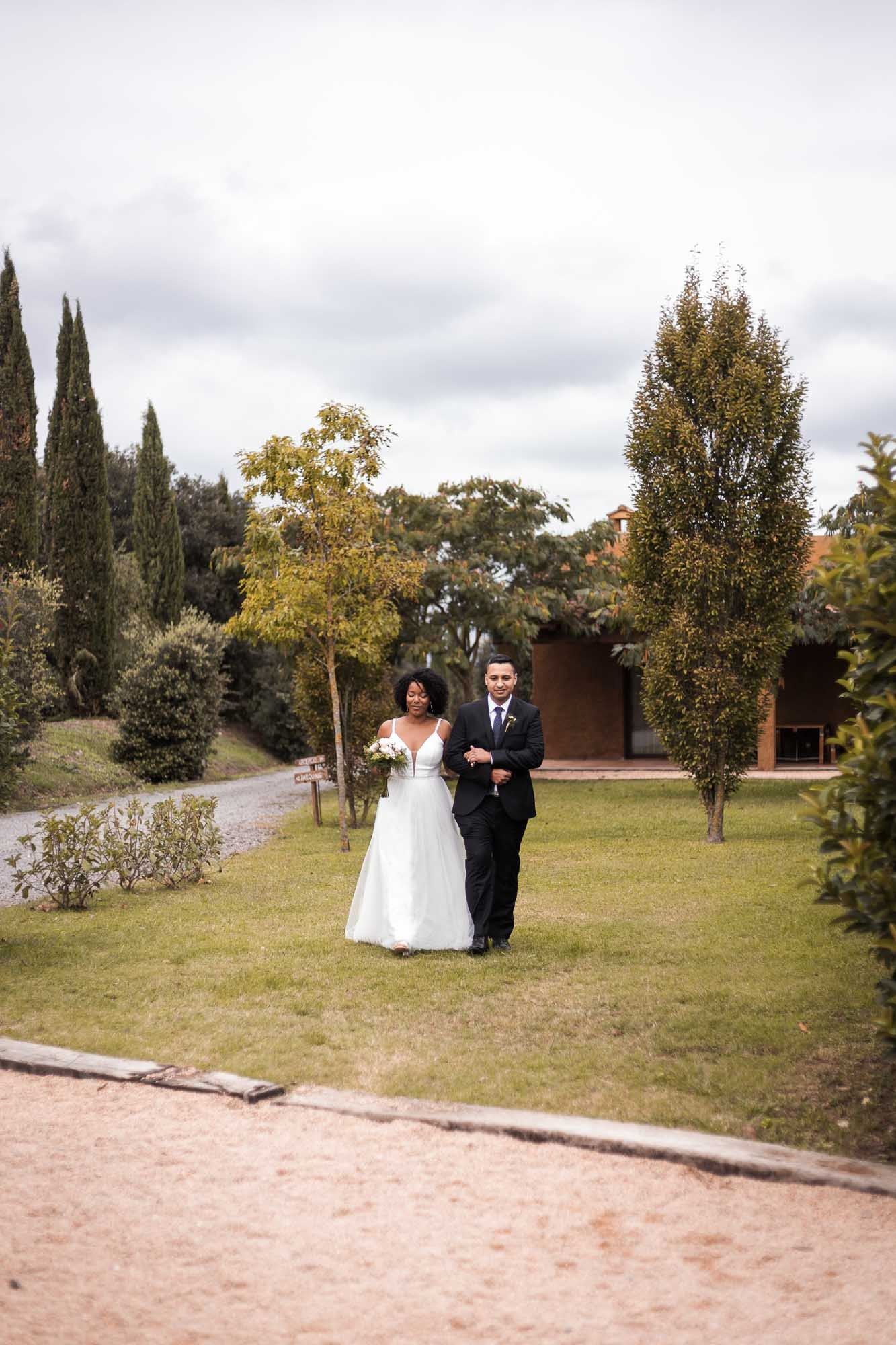The couple stand small in a wide garden with a gravel path and tall cypress trees under overcast light, Mariana and Arnau’s wedding at La Vaqueria de Mas Garriga