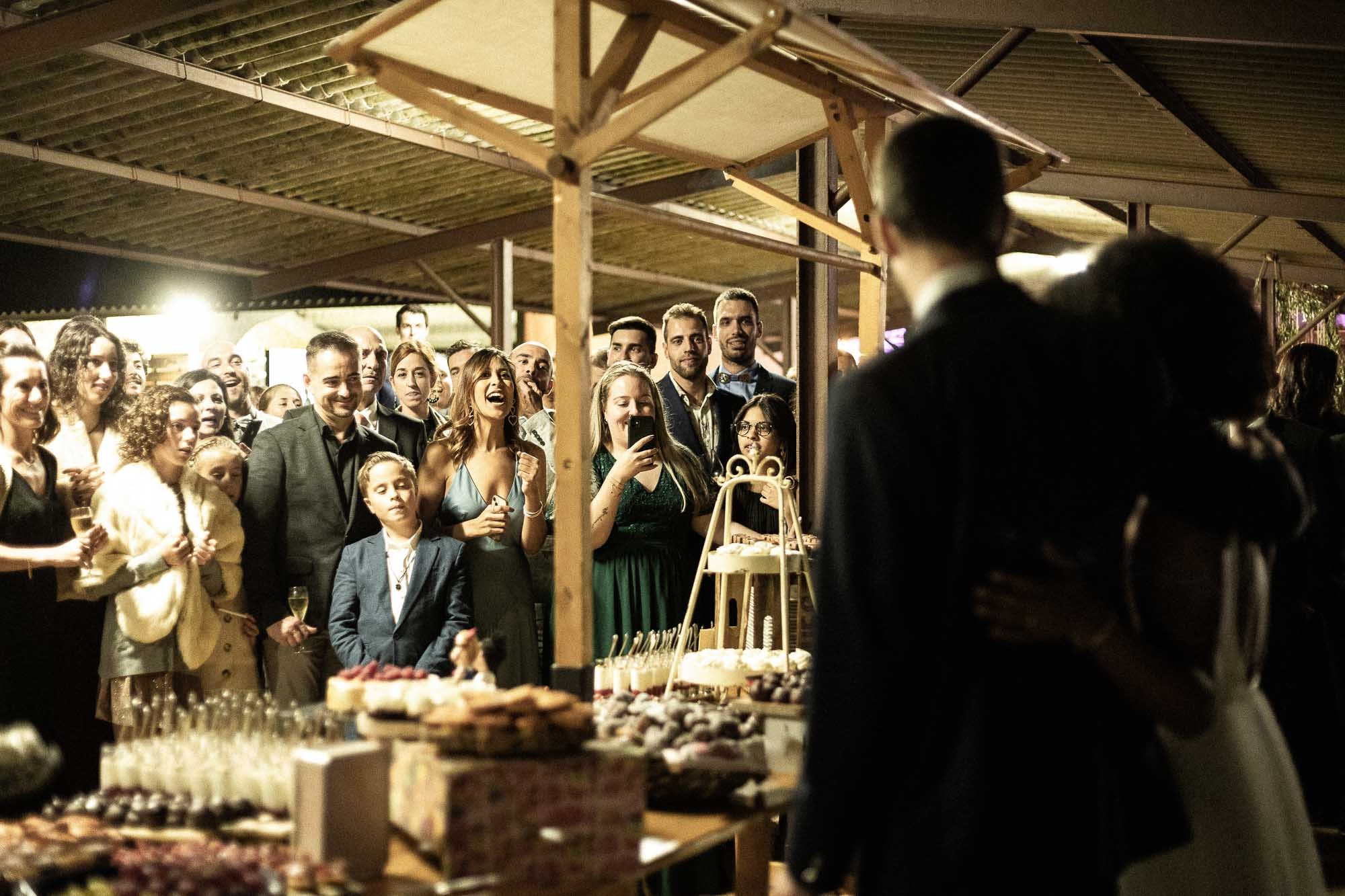 Invités réunis autour d’une longue table de nourriture sous les poutres en bois de La Vaqueria de Mas Garriga, avec Mariana et Arnau au premier plan