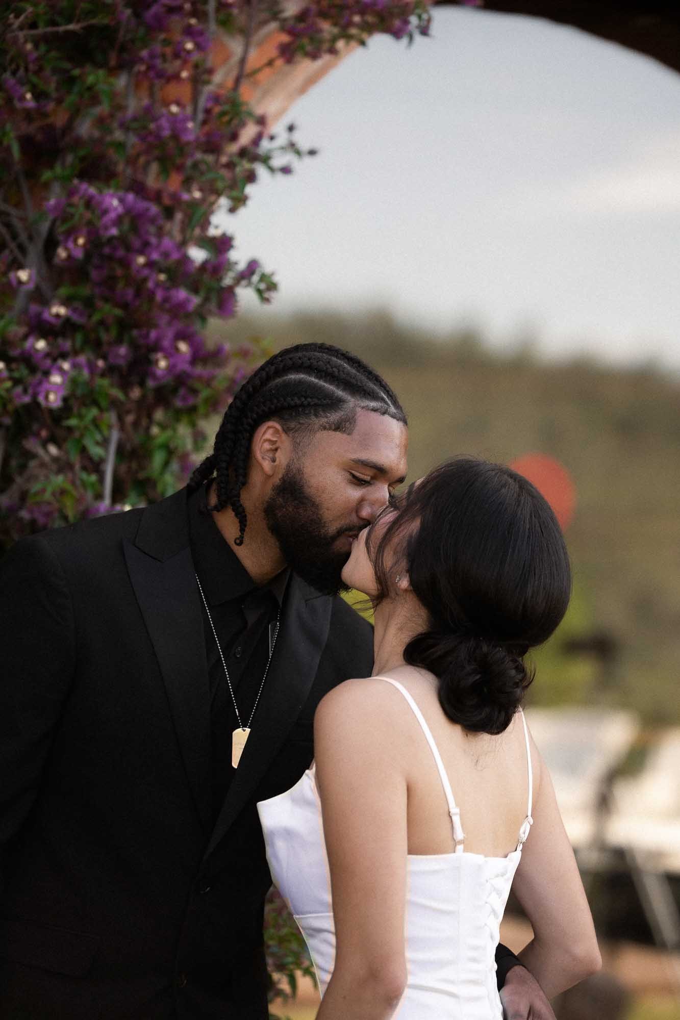 The couple kiss at the ceremony beside purple flowers and a brick arch, hills visible behind, Mercedes and Jay’s wedding at Can Borrell, near Barcelona