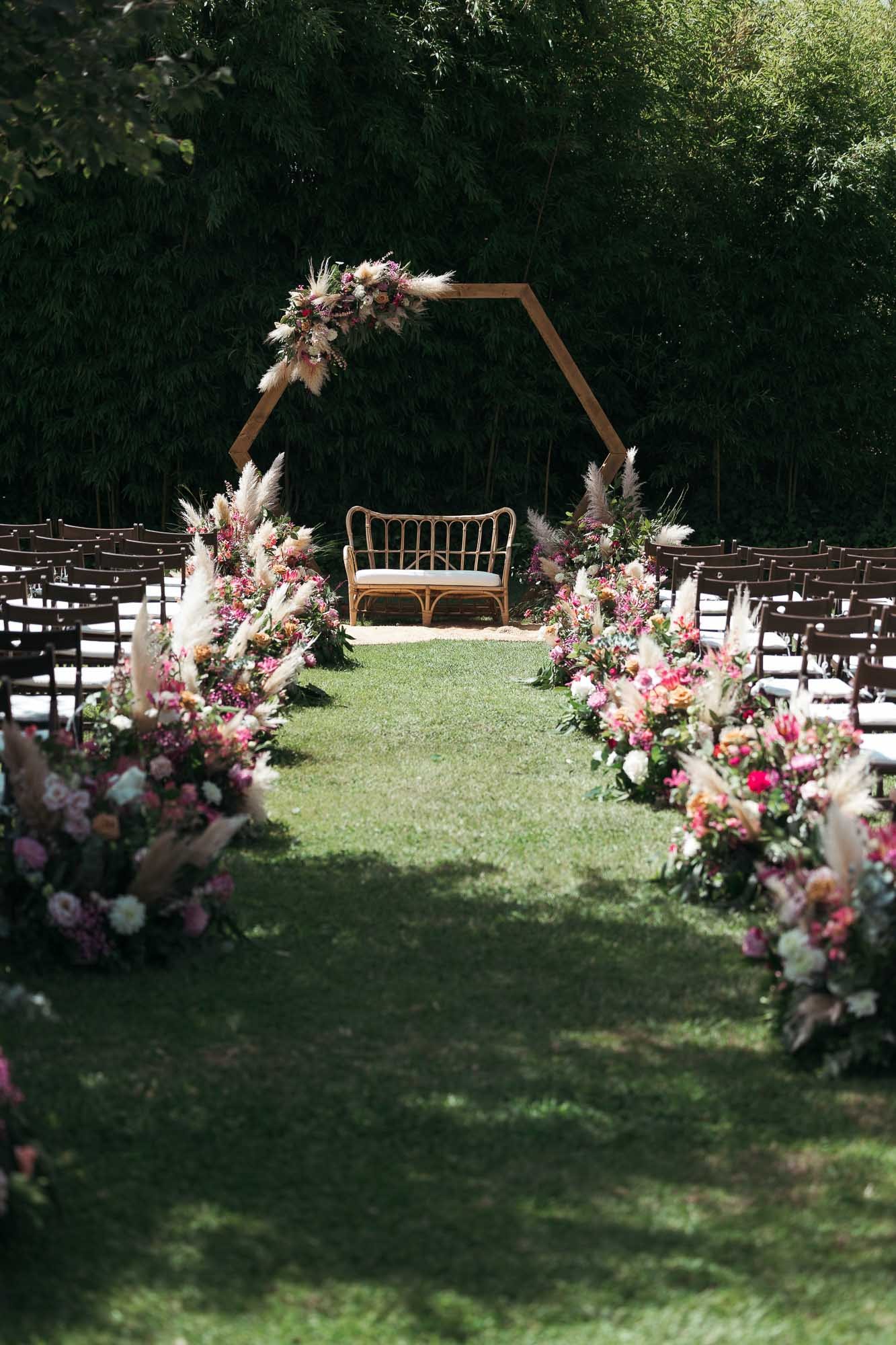 Grass aisle strewn with petals between rows of chairs leading to a hexagonal wooden floral arch in the garden, Edu and Míriam’s wedding at Mas Palau, Costa Brava