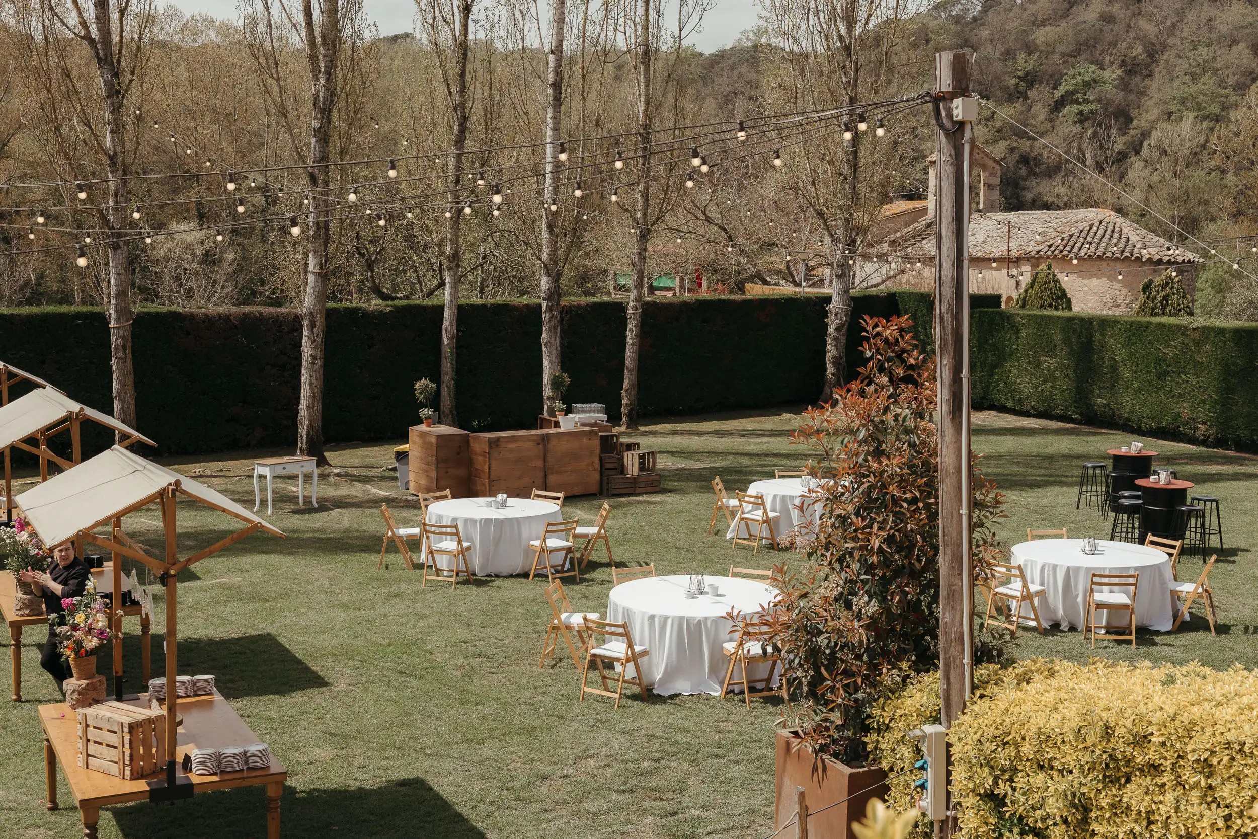 Vue large du jardin d’El Jardí de Calders avec des tables rondes sous de grands arbres et des guirlandes lumineuses, avant l’arrivée des invités