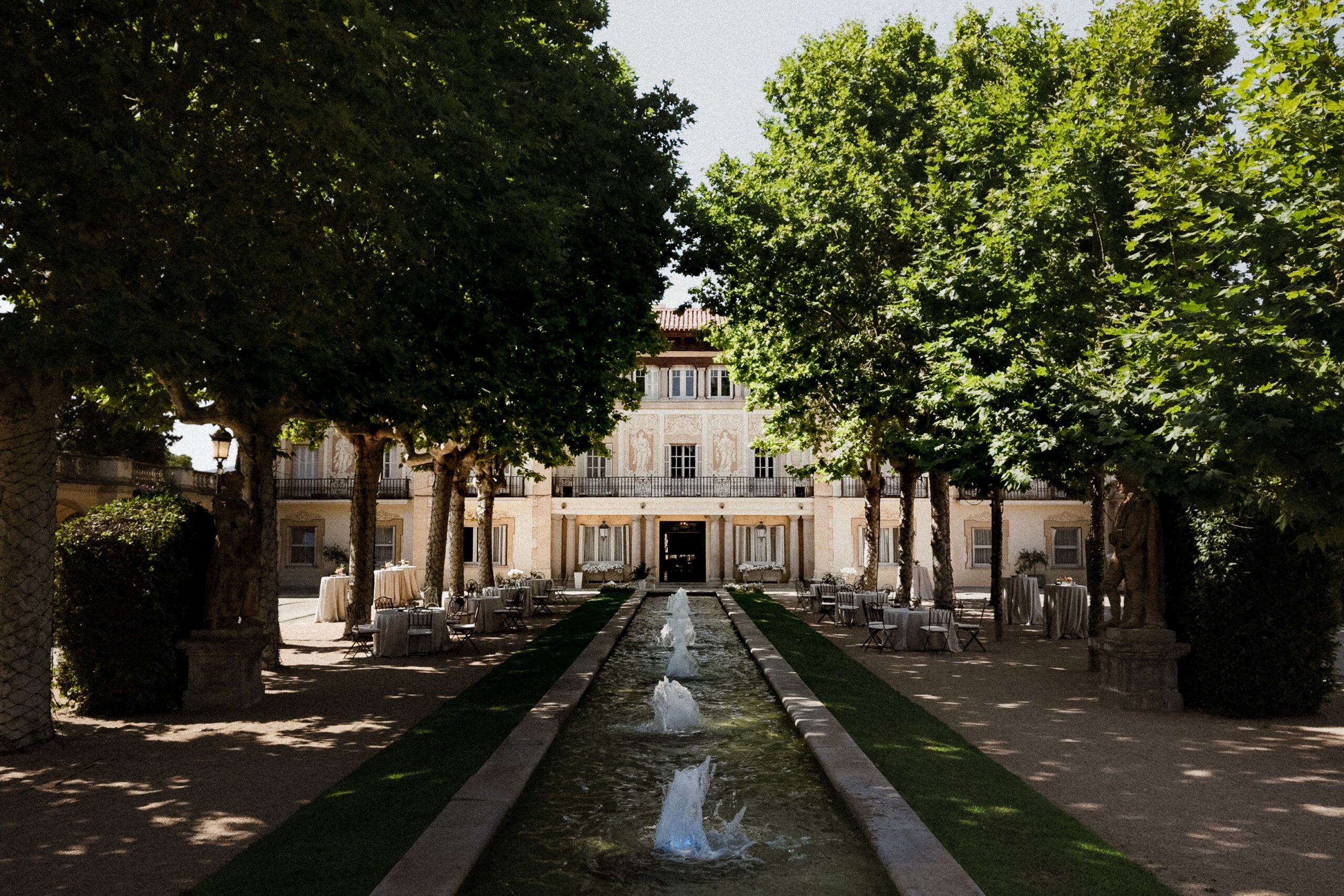 Symmetrical view down a water channel between tall trees toward a stone villa facade in dappled sunlight, wedding photography in Catalonia