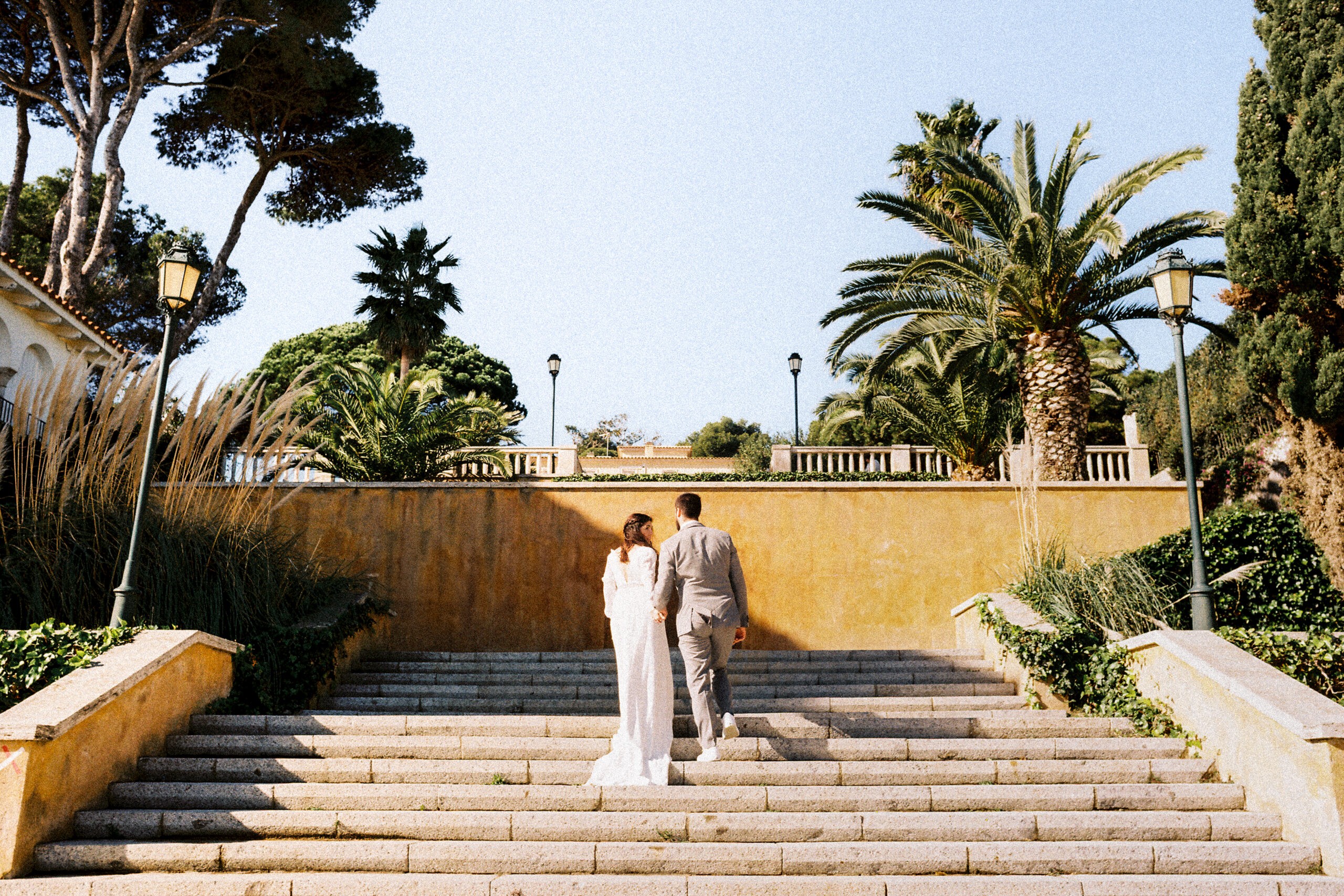 The couple walk up a wide stone staircase flanked by palm trees under a clear blue sky, wedding photography in Catalonia