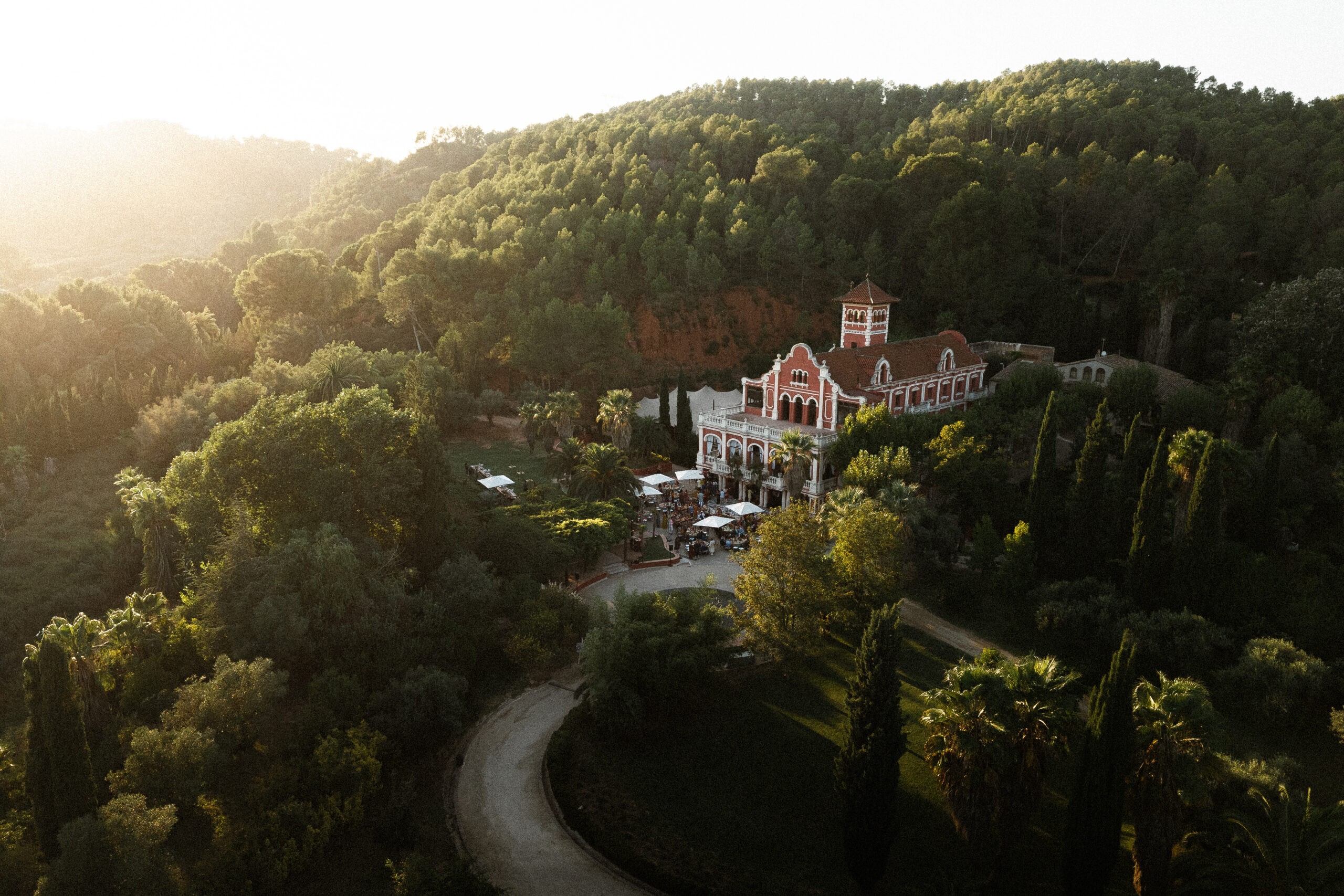 Aerial view of forested hills and a manor house at golden hour, a winding road below, wedding photography in Catalonia