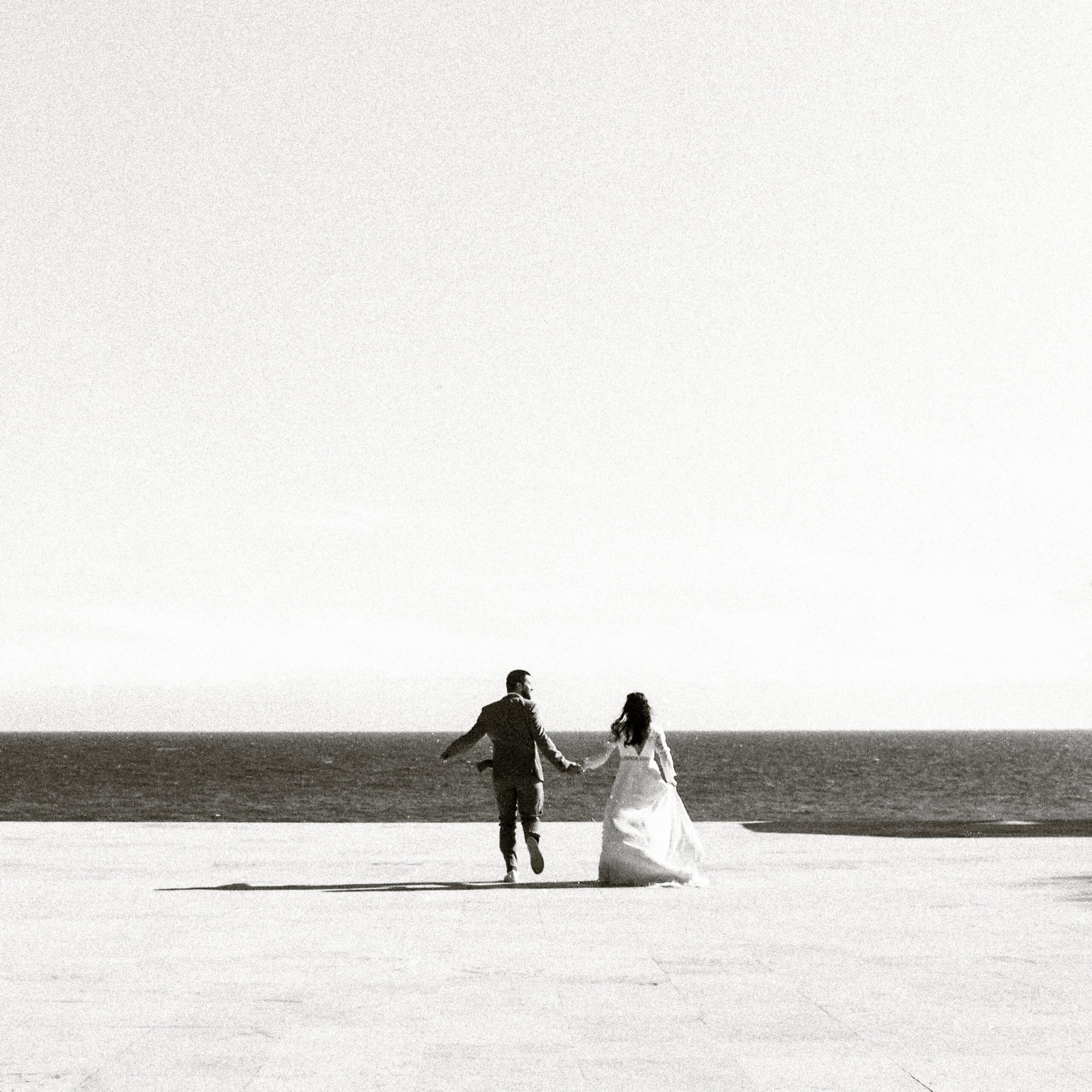 Photographie en noir et blanc d’un couple qui marche main dans la main vers la mer sur une promenade claire et lumineuse, issue d’une de nos séances de portraits de mariage.