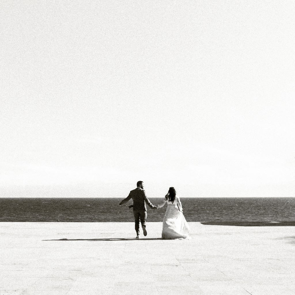 Black and white shot of the couple walking hand in hand toward the sea across a pale, bright promenade, wedding photography in Catalonia