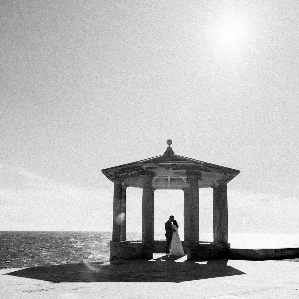 Black and white shot of the couple small beneath a stone pavilion by the sea, sun flaring above, wedding photography in Catalonia
