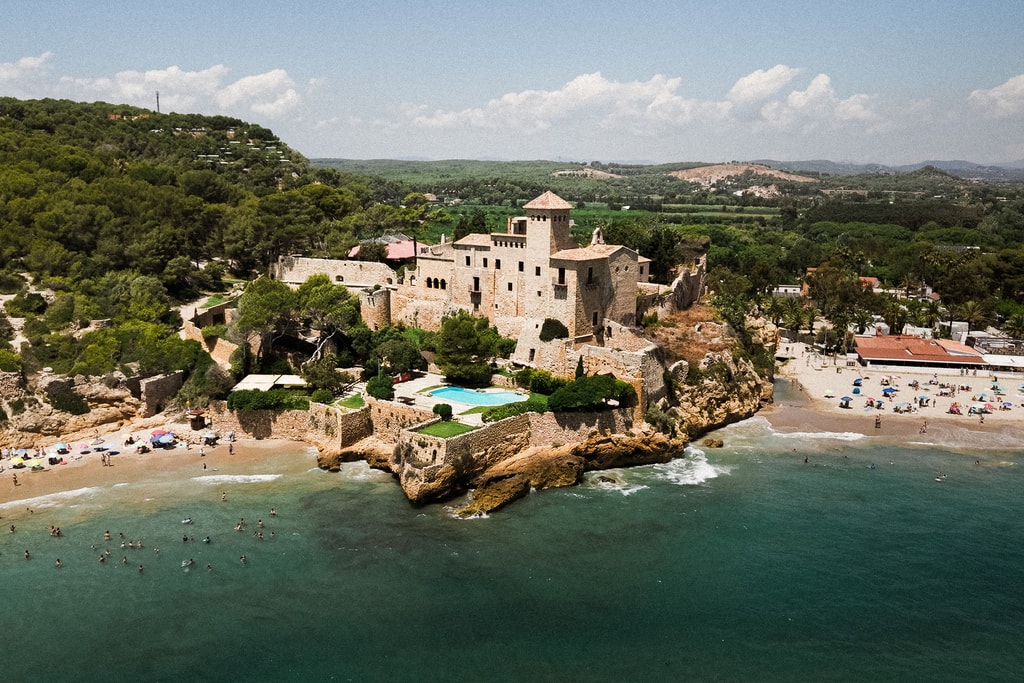 Aerial view of a castle-like estate on a rocky headland beside a sandy cove and turquoise sea, wedding photography in Catalonia