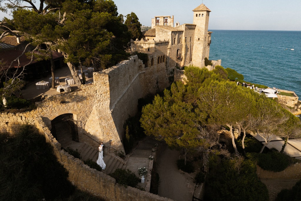 Aerial view of a castle on a cliff above the sea with stone walls, pine trees and a garden path, wedding photography in Catalonia