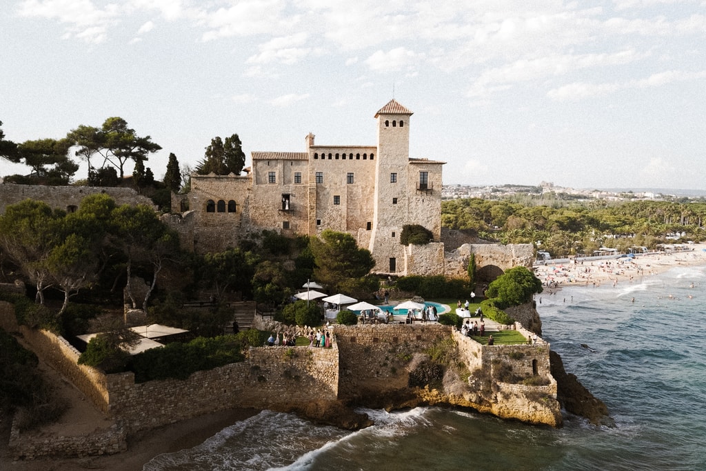 Aerial view of a castle on a rocky headland above the sea, with a beach and pine trees beside it, wedding photography in Catalonia