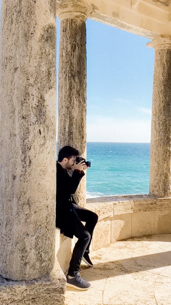 Photographer sitting between stone columns with a camera and a view of the sea