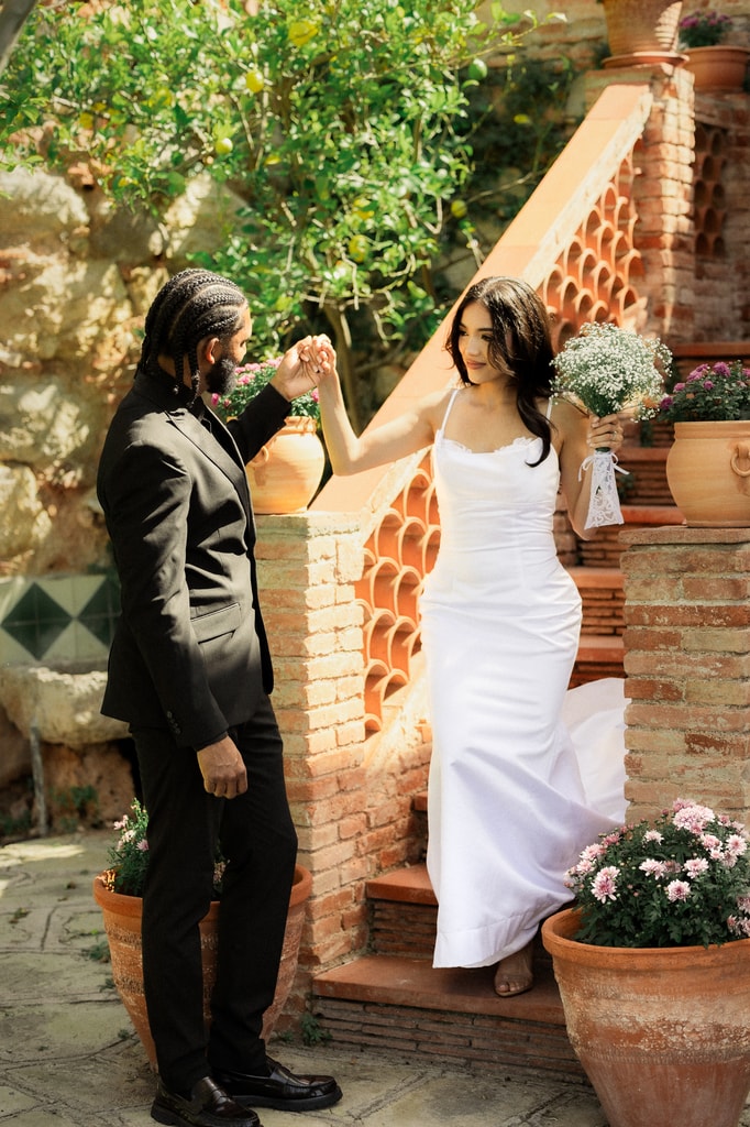 The groom in black helps the bride in a white satin dress down brick steps among terracotta pots, wedding photography in Catalonia