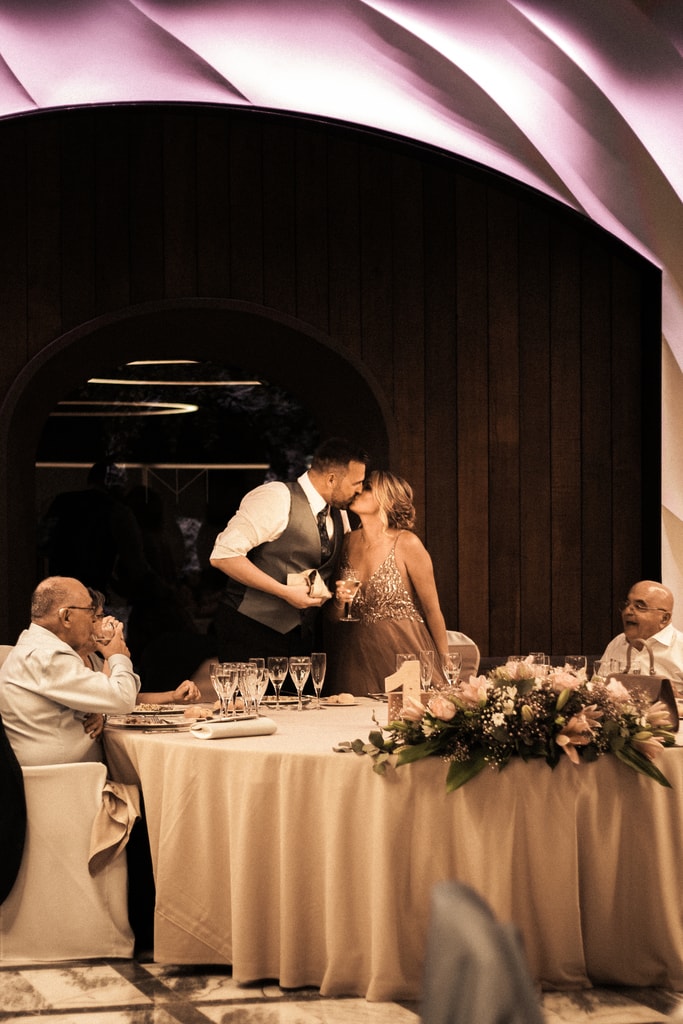The couple kiss at the head table in a hall with a pink sculpted ceiling and dark wooden walls, wedding photography in Catalonia