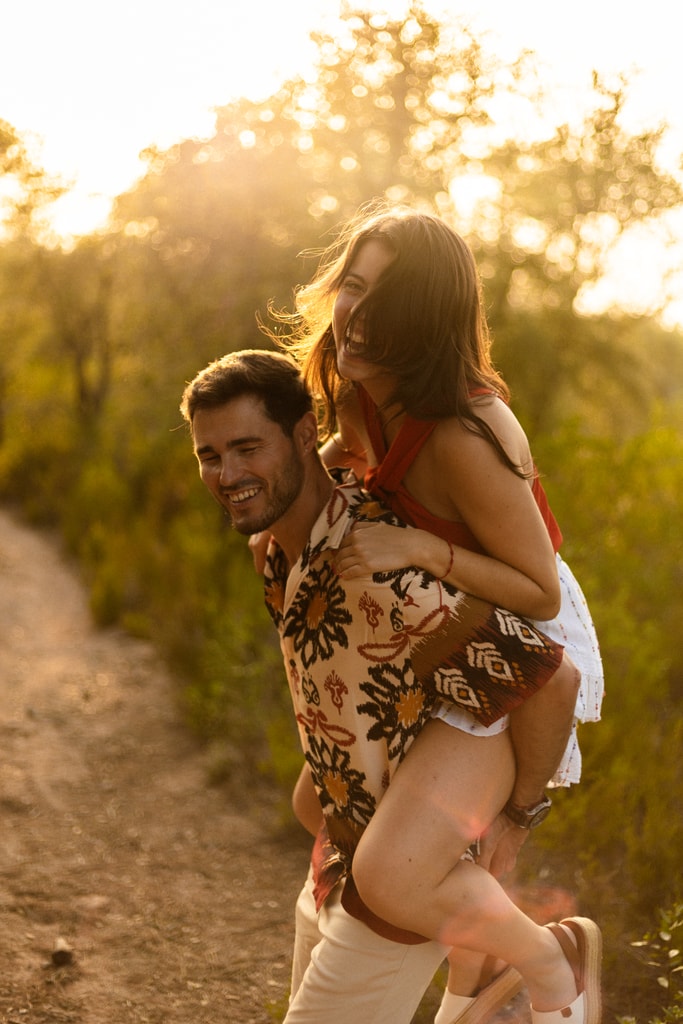 A man carries a laughing woman piggyback along a dirt path at golden hour, shrubs behind them, wedding photography in Catalonia