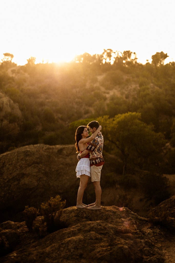 The couple embrace on a rock at sunset above forested hills, golden light, wedding photography in Catalonia