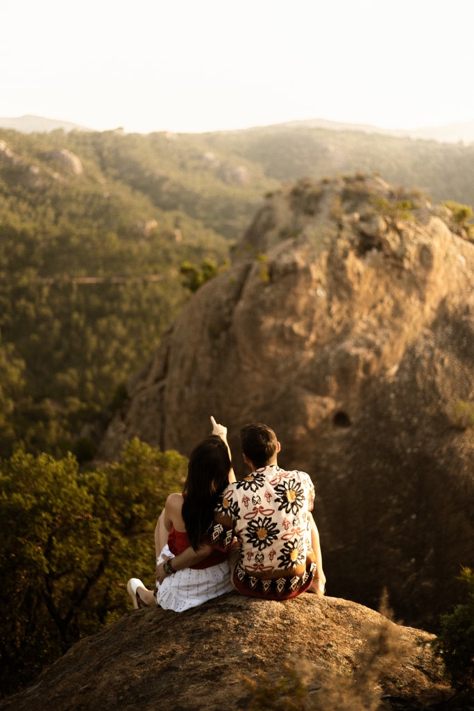 The couple sit side by side on a rock looking at forested hills lit by golden light, wedding photography in Catalonia