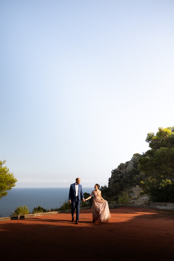 The couple stand small on a headland above the sea under a pale blue sky, wedding photography in Catalonia