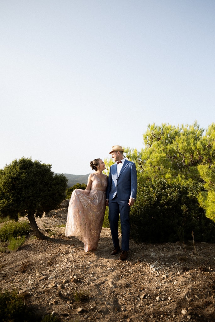 The couple stand on a hillside among trees, the groom in a navy suit and hat, the bride in a pink dress, wedding photography in Catalonia
