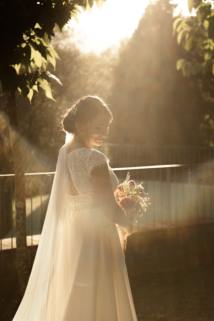 The bride in a long dress and cape turns away, backlit by sun through trees, bouquet in hand, wedding photography in Catalonia