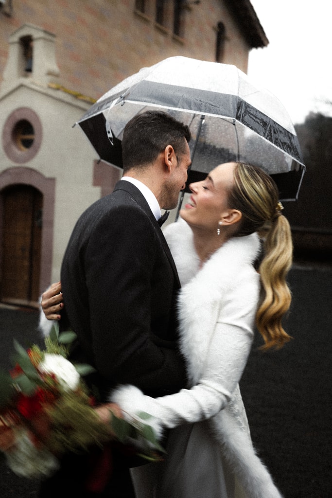 The couple embrace under a clear umbrella in front of a small chapel, the bride in a white coat with a bouquet, wedding photography in Catalonia