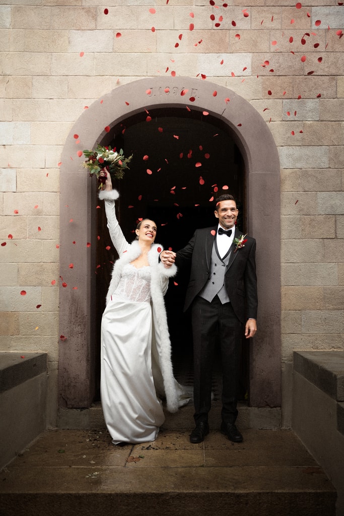 The couple step out of a stone arched doorway as red petals rain down, the bride raising her bouquet, wedding photography in Catalonia