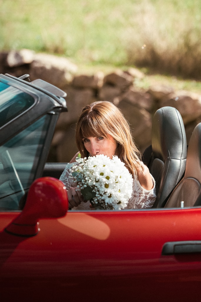 The bride sits in a red convertible looking down at her bouquet of white flowers, green foliage behind, wedding photography in Catalonia