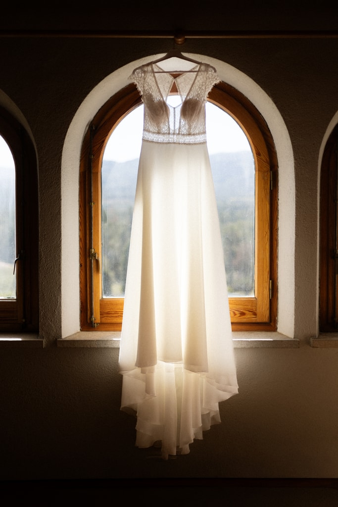 Wedding dress hanging in an arched window with backlight and a view of mountains beyond, wedding photography in Catalonia