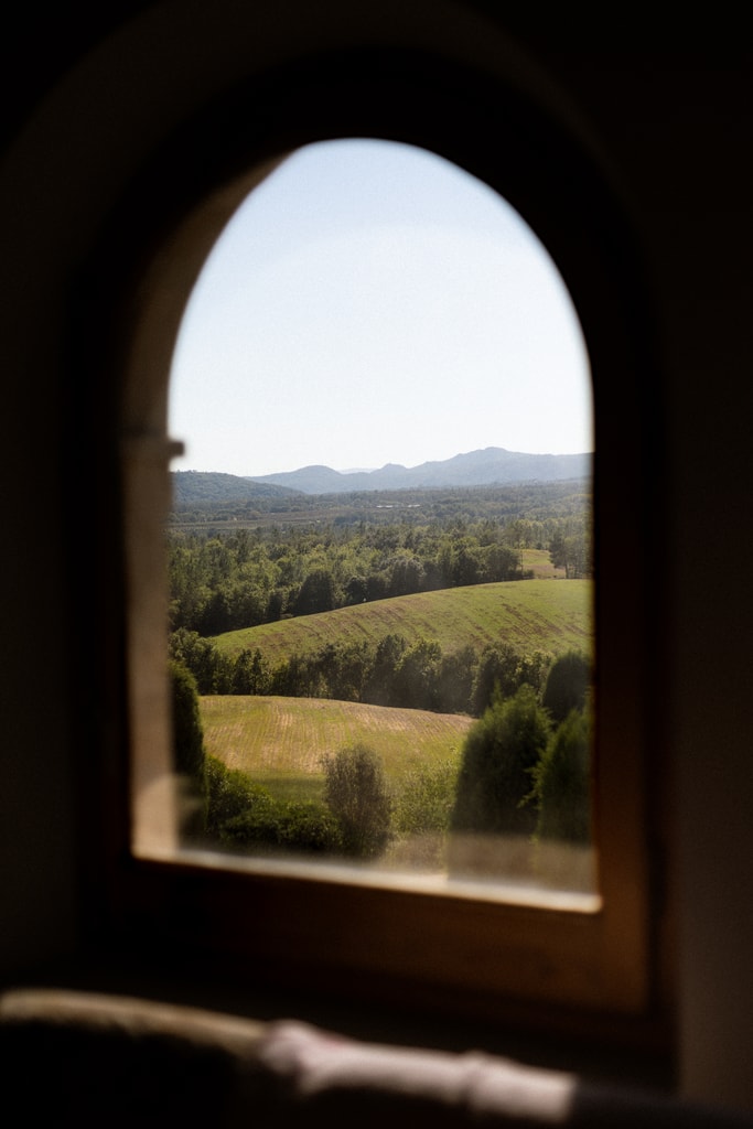 Arched window framing rolling green hills and a distant mountain range from a dark interior, wedding photography in Catalonia
