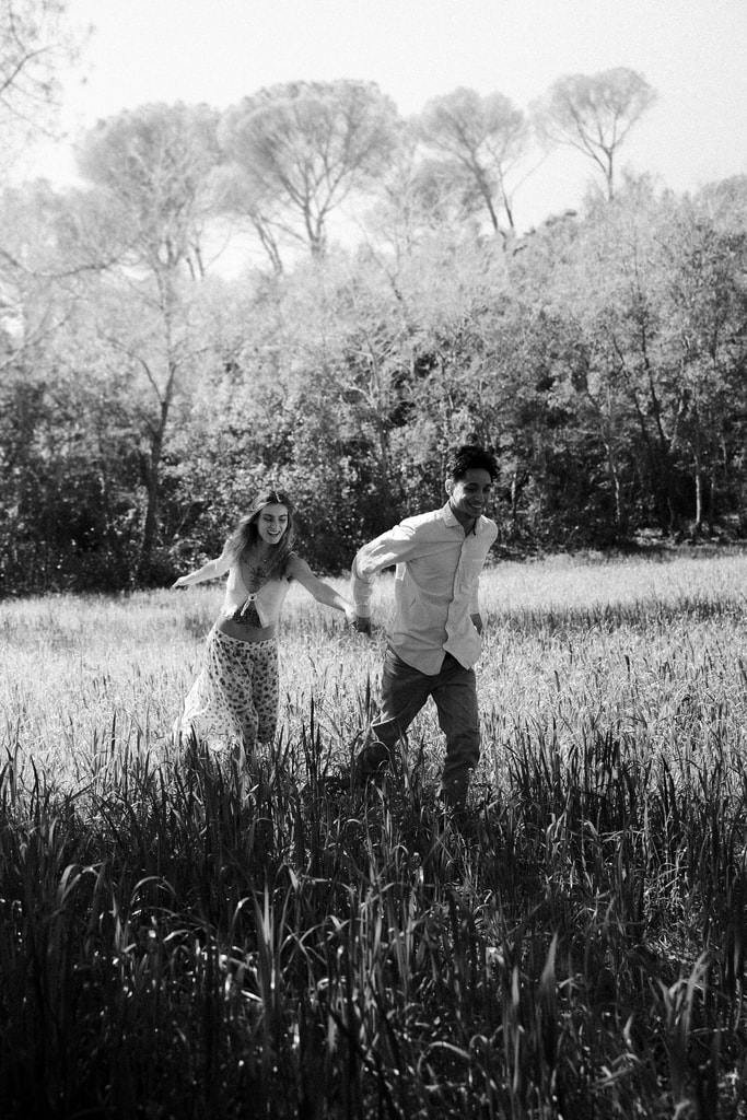 Black and white shot of the couple walking through tall grass with pine trees behind them, wedding photography in Catalonia