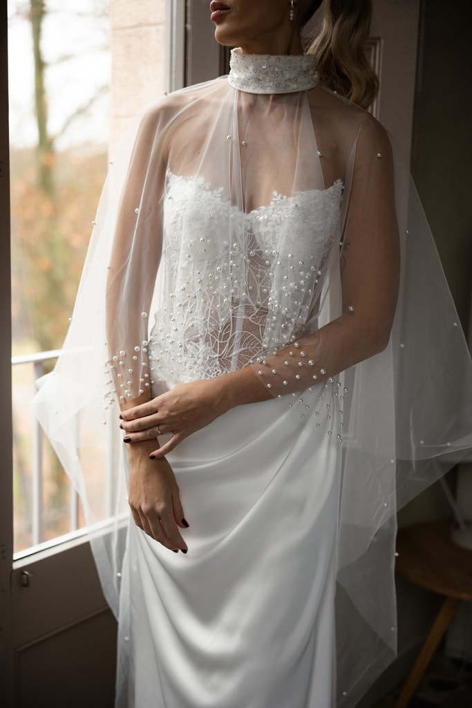 The bride in a lace gown with a sheer high-neck cape stands by a bright window, face cropped out, wedding photography in Catalonia