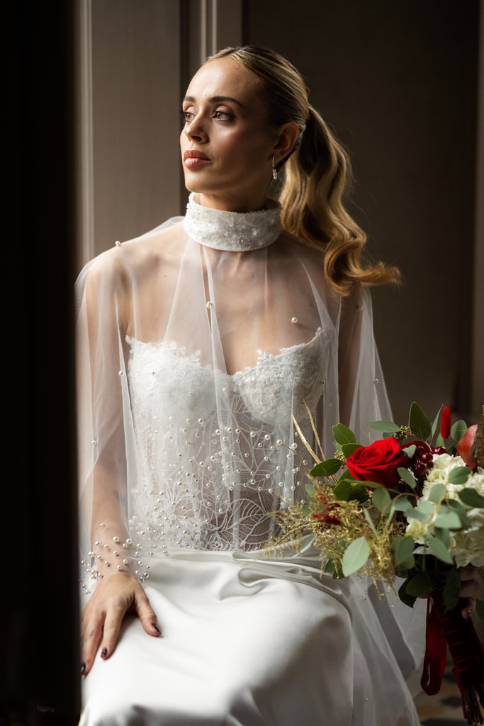 The bride with long wavy hair and a sheer overlay sits with a red and white bouquet against a dark background, wedding photography in Catalonia