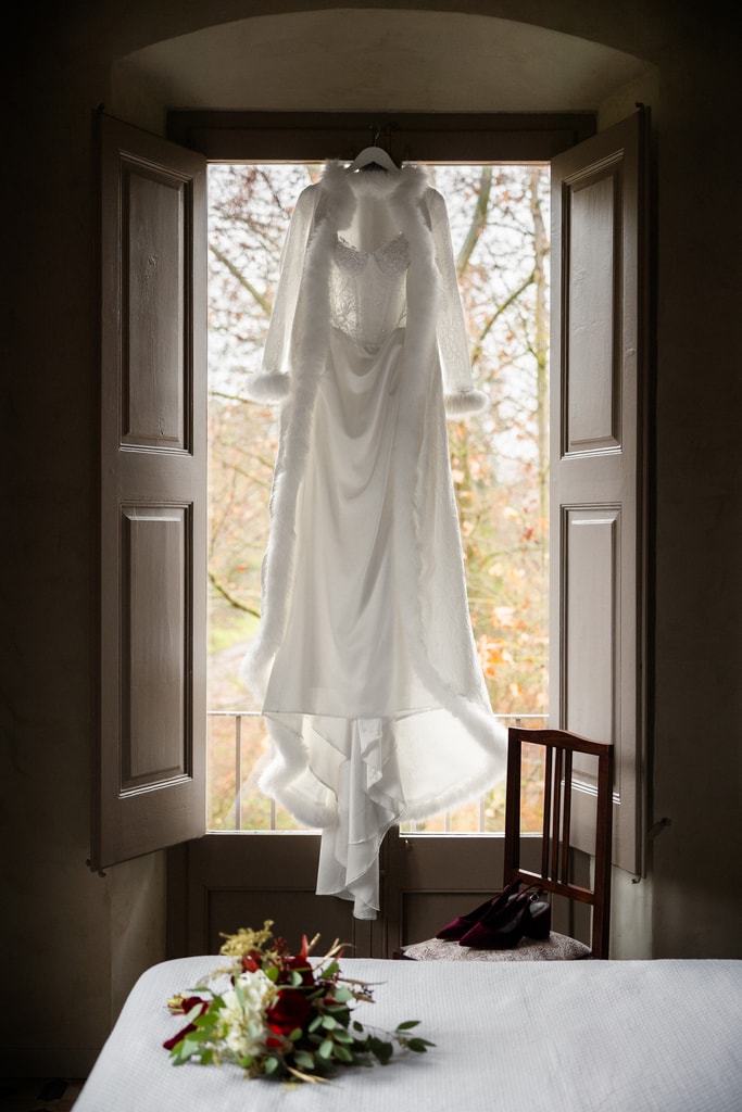 Wedding dress hanging in a tall shuttered window with a bouquet on a white table below, wedding photography in Catalonia