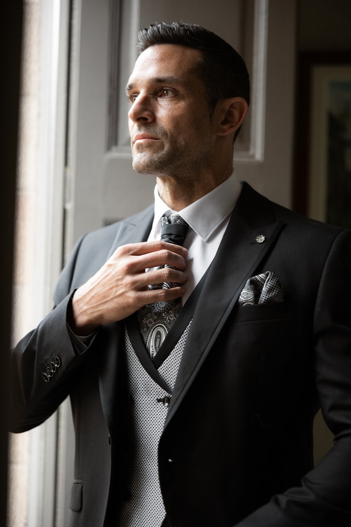 The groom in a dark three-piece suit adjusts his tie beside a window, looking aside in soft light, wedding photography in Catalonia