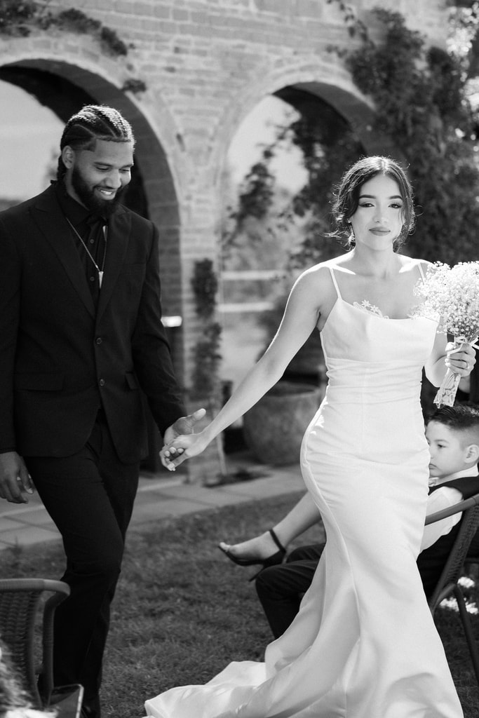 Black and white shot of the couple walking hand in hand and laughing, brick arches behind them, wedding photography in Catalonia