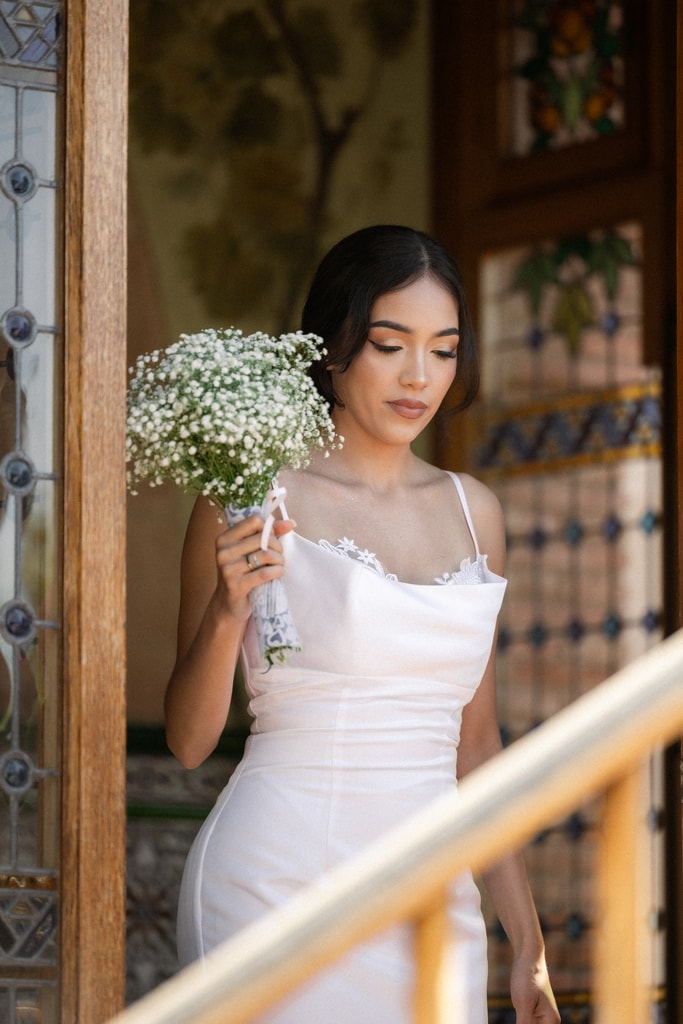 The bride in a white satin dress holds a baby’s breath bouquet beside a stair rail and stained glass, wedding photography in Catalonia