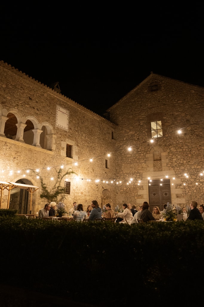 Stone courtyard at night with an arched gallery, string lights and guests dining below, wedding photography in Catalonia