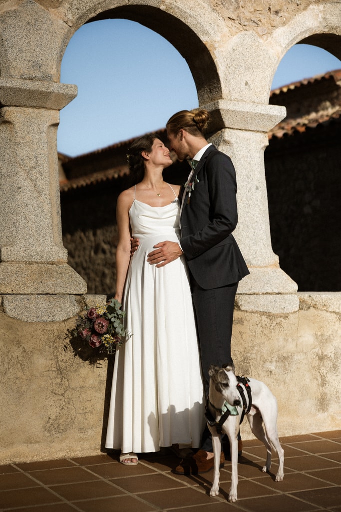 The couple kiss beneath stone arches while a greyhound stands at their feet, warm light, wedding photography in Catalonia