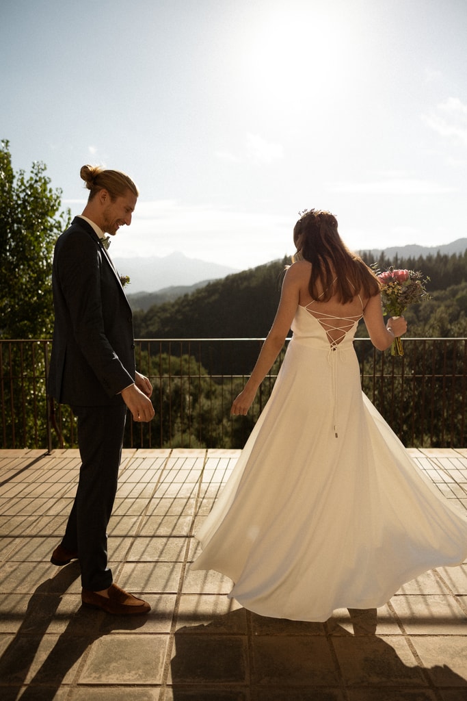 The groom looks at the bride as her dress swirls on a wooden terrace with mountains behind, low sun, wedding photography in Catalonia