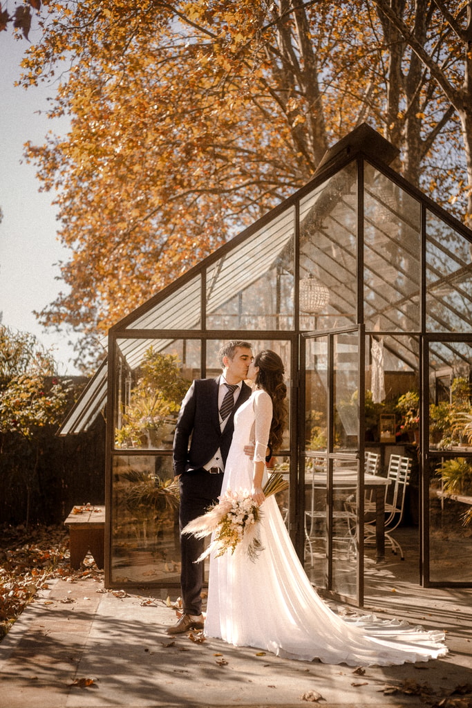 The couple embrace inside a glasshouse surrounded by orange autumn trees, the bride holding a bouquet, wedding photography in Catalonia