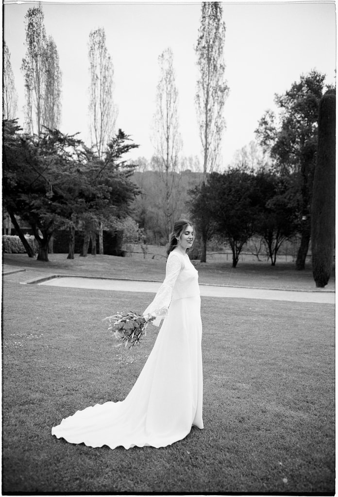 Black and white shot of the bride in a long dress holding a bouquet on a lawn, tall poplars behind, wedding photography in Catalonia