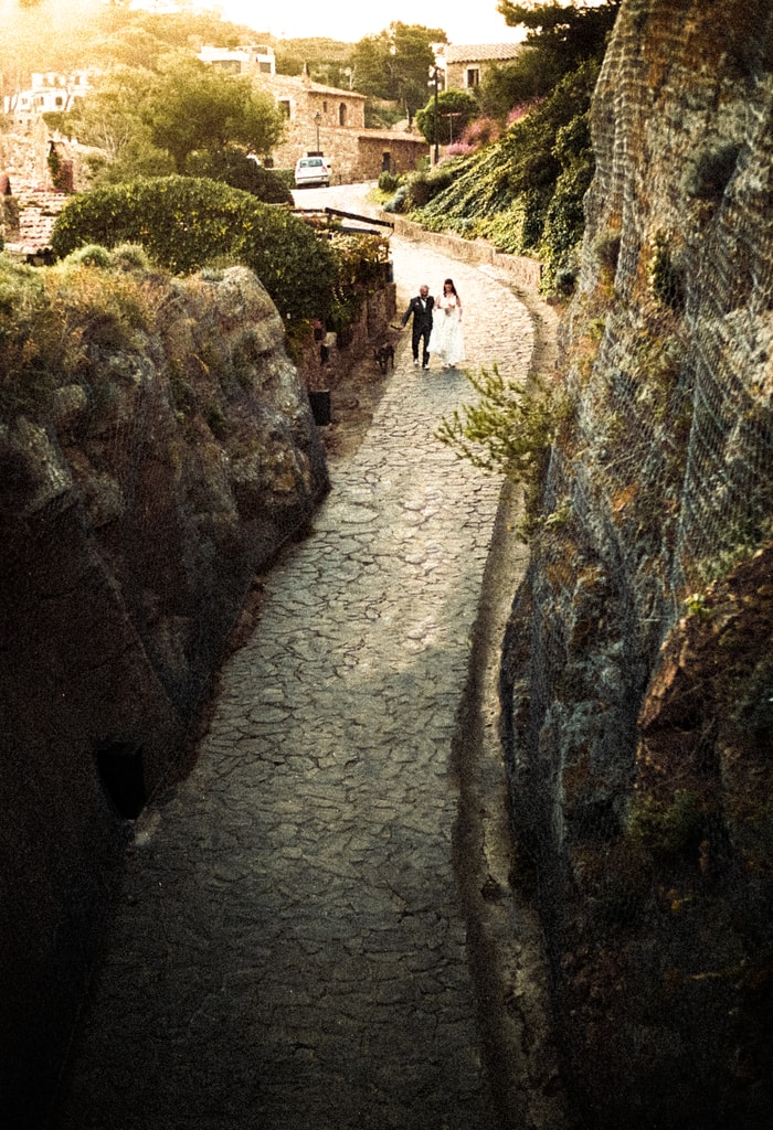 Narrow stone path between rocky walls in golden light, a couple walking small in the distance, wedding photography in Catalonia