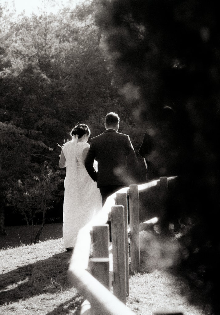 Black and white shot of the couple walking away along a wooden railing under trees, bride in a long dress, wedding photography in Catalonia