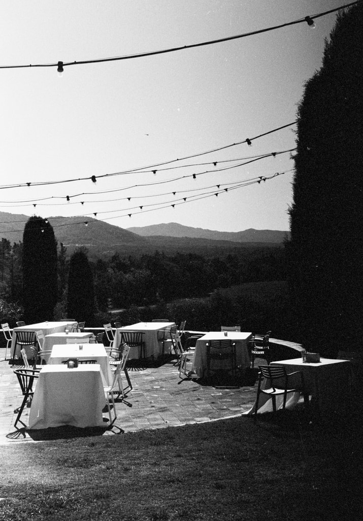 Black and white view of a terrace with set round tables and string lights, hills in the distance, wedding photography in Catalonia