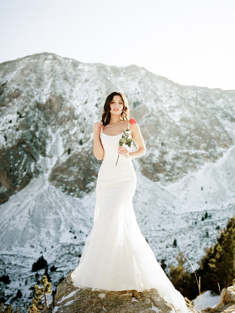 The bride in a fitted white gown stands on a pale rocky mountain slope, bouquet in hand, looking aside, wedding photography in Catalonia