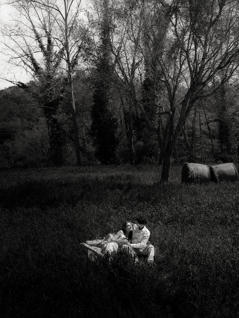 Black and white shot of a couple lying on the grass in dark woods, trees bare behind them, wedding photography in Catalonia